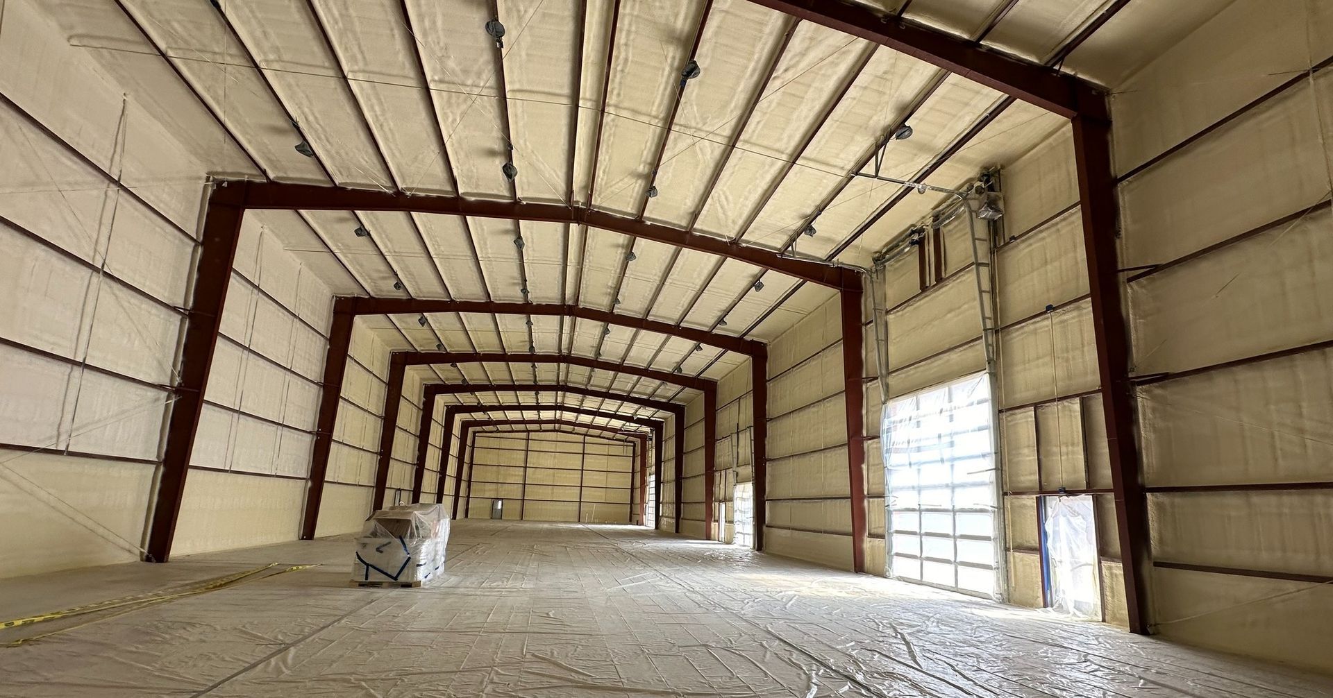 Empty industrial warehouse interior with exposed metal beams and concrete floor, lit by daylight from large windows
