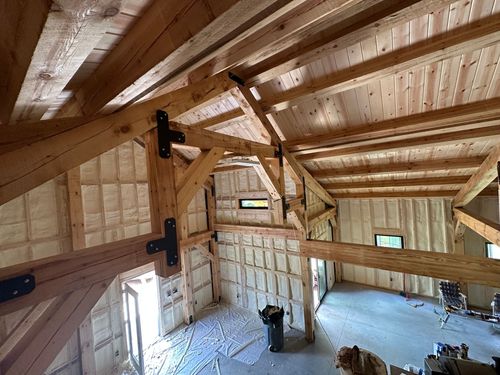 Interior of a wooden loft with exposed beams, wall panels, and small windows under a sloped roof.