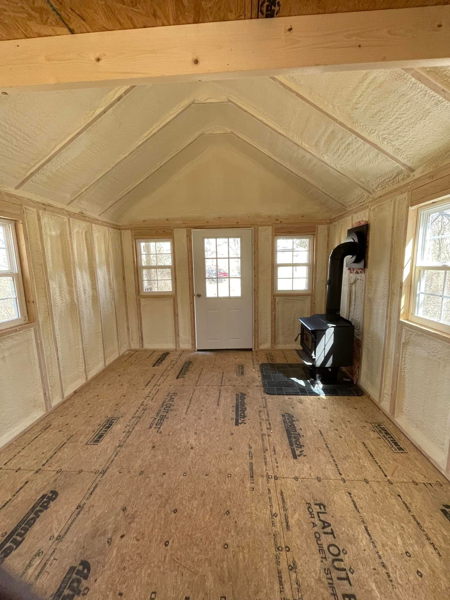 Unfinished small cabin interior with insulated walls, OSB floor, windows, a door, and a wood stove.