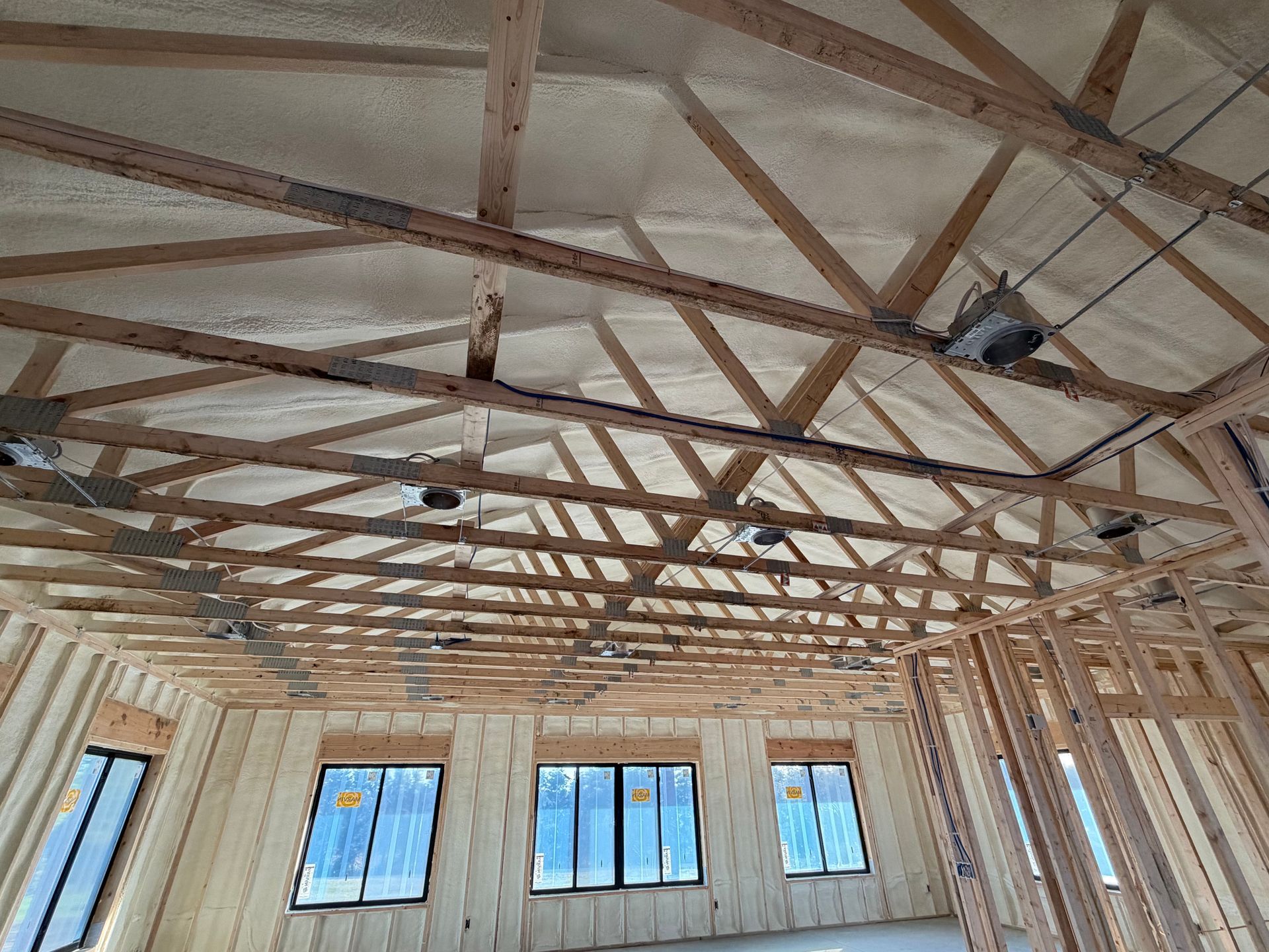 Interior of a wooden-framed building under construction with exposed roof trusses and several windows along the wall