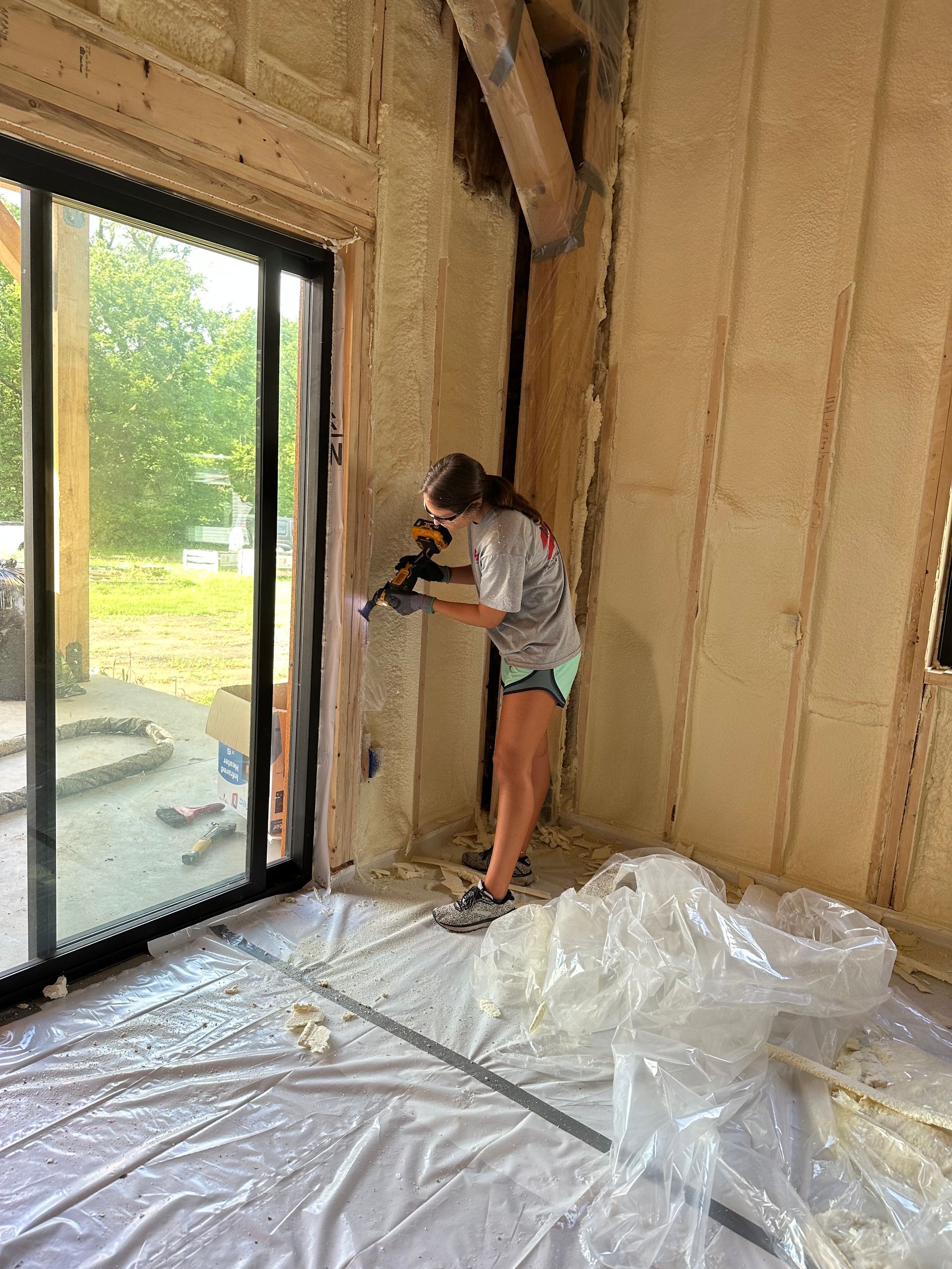 Person painting a beige room wall beside a sliding glass door, with plastic sheeting covering the floor.